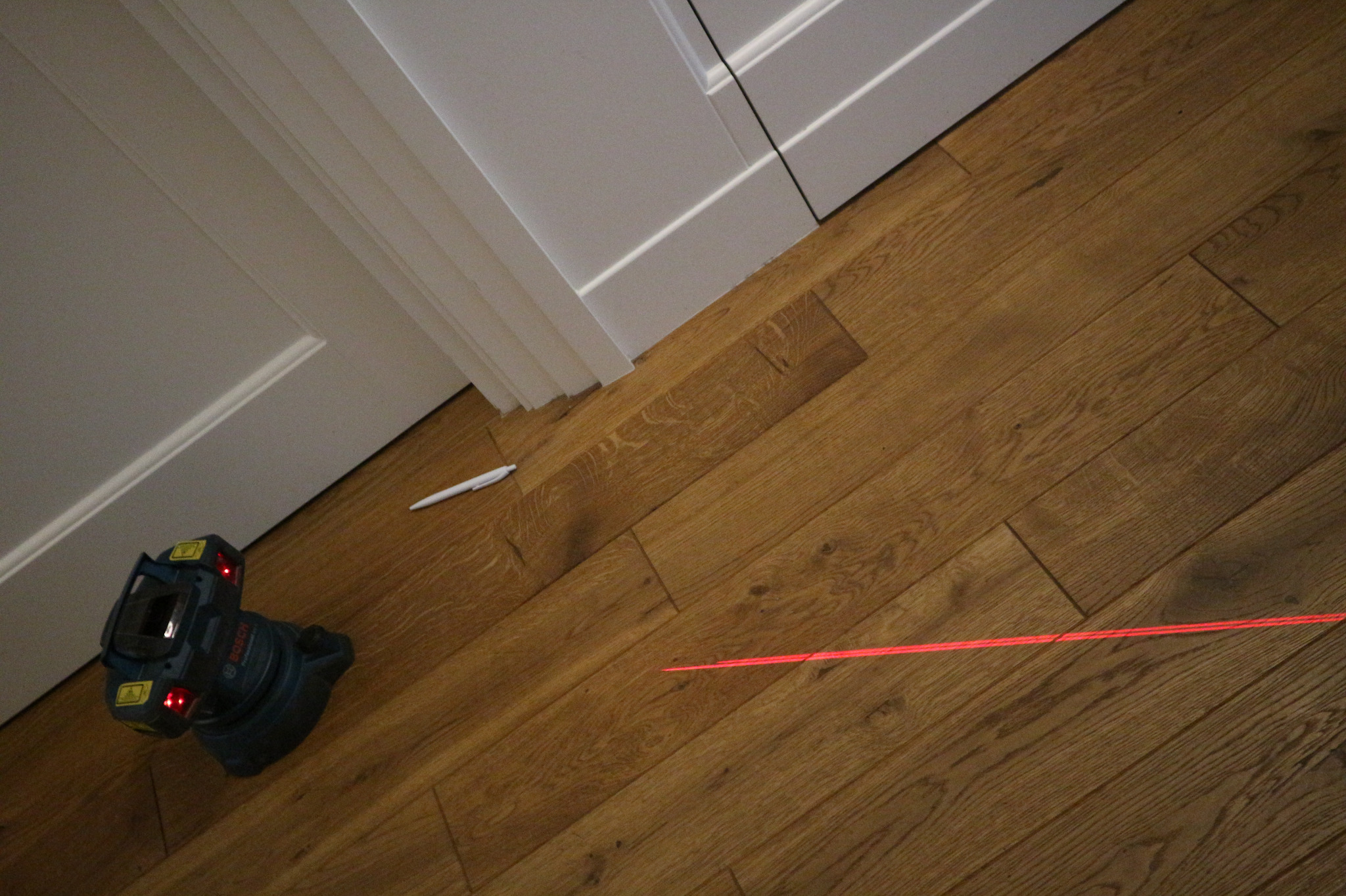 Poziomica laserowa Bosch rzuca czerwoną linię na parkiet z desek, obok biały ołówek i fragment białej listwy przypodłogowej.