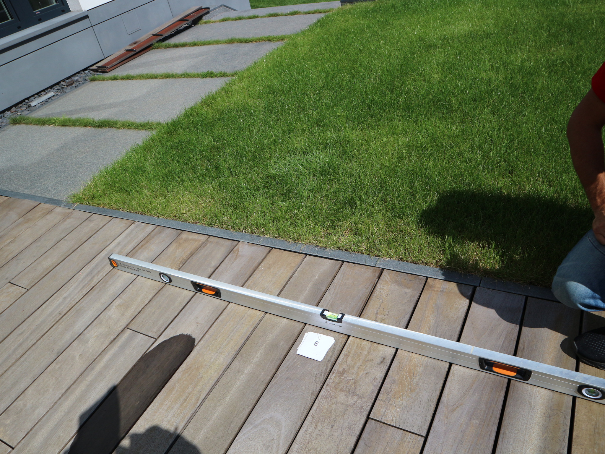 Poziomnica na drewnianym tarasie, fragment trawnika i kostki brukowej w tle, widoczny cień osoby.
