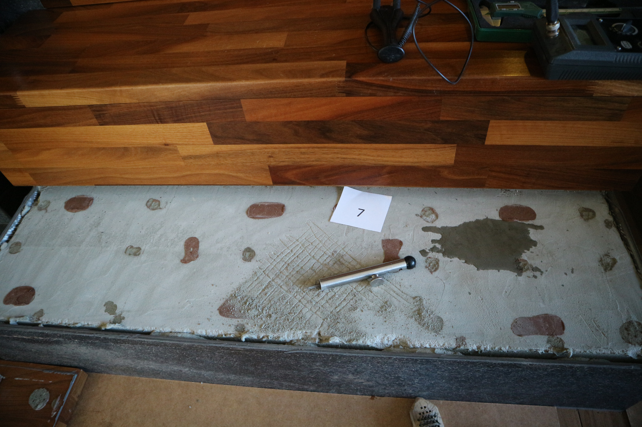 Betonowa płyta z widocznymi śladami po badaniach, narzędziem pomiarowym i kartką z numerem 7, umieszczona pod drewnianym blatem.