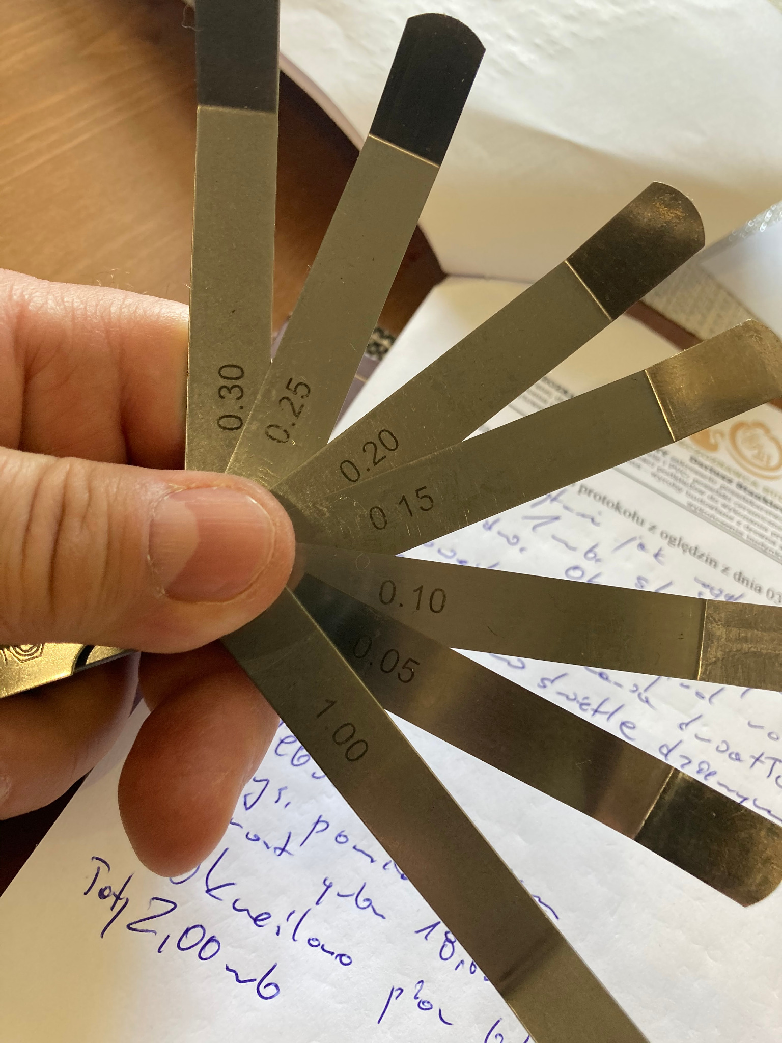 Zestaw szczelinomierzy w dłoni, z widocznymi wartościami grubości od 0.05 do 0.30 mm, leżący na dokumencie z odręcznymi notatkami.