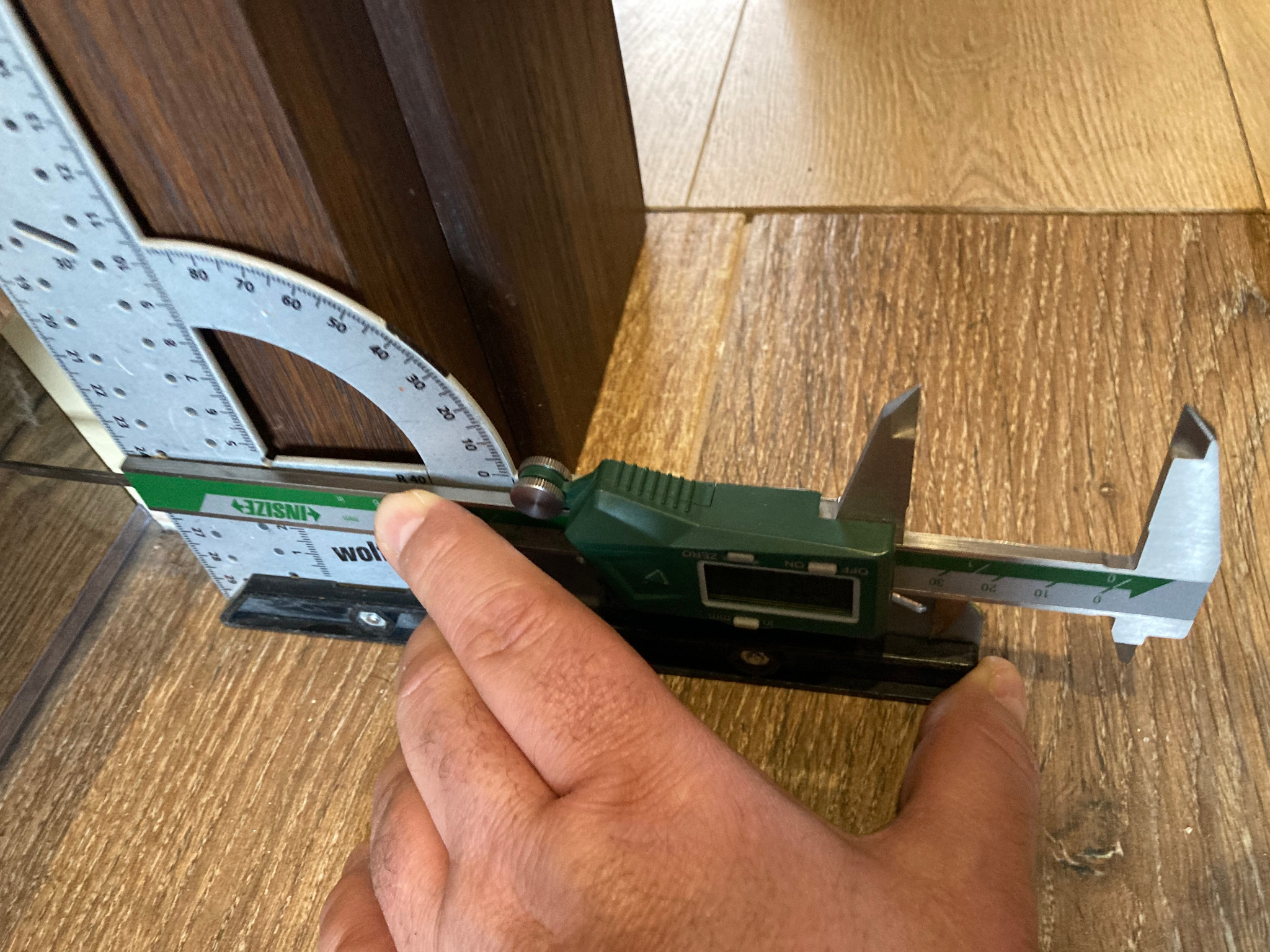Pomiar grubości drewnianego elementu za pomocą suwmiarki cyfrowej Insize z kątomierzem, na tle drewnianej podłogi.