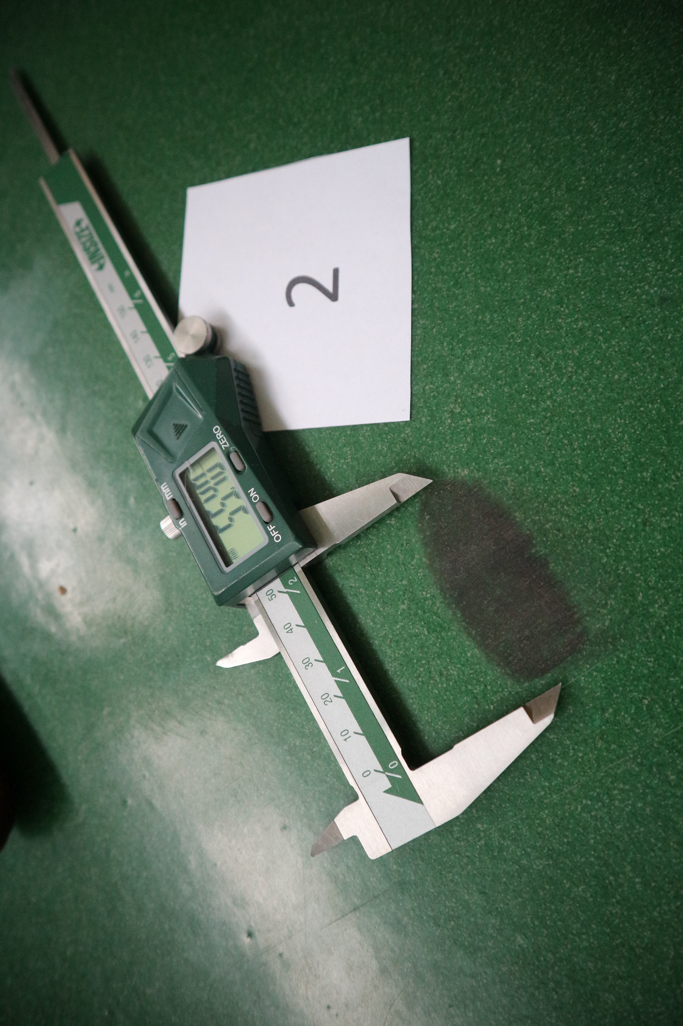 Elektroniczna suwmiarka INSIZE wyświetlająca pomiar 55.48 mm, leżąca na zielonym tle obok kartki z numerem 2.