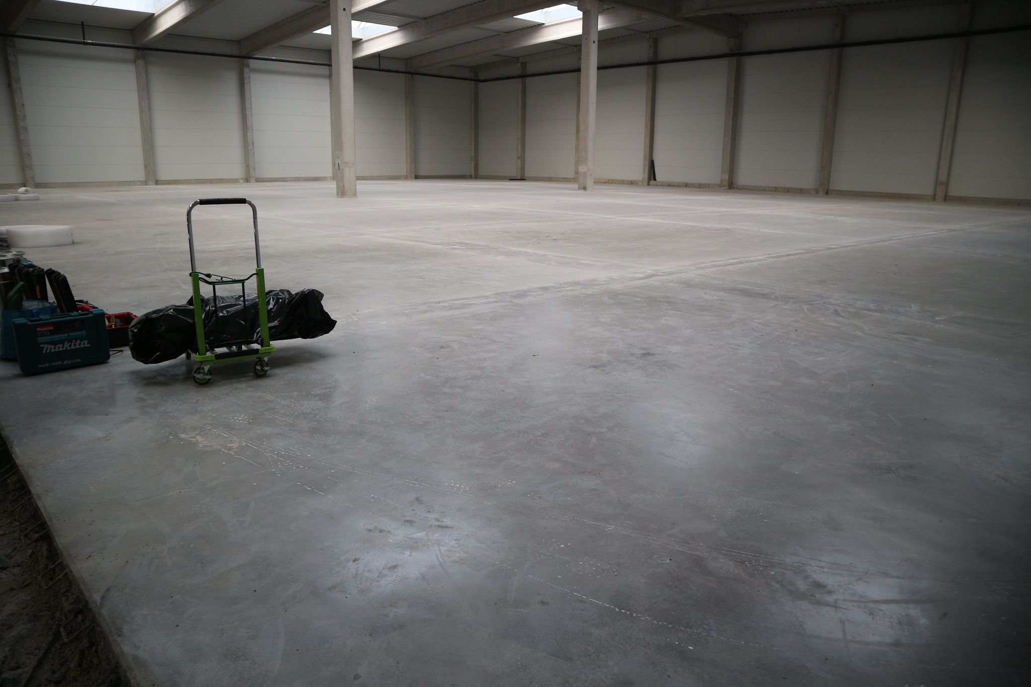 Wnętrze pustej hali magazynowej z betonową posadzką, w rogu wózek transportowy z ładunkiem pod czarną folią i skrzynka narzędziowa Makita.