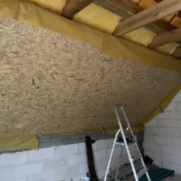 Ocieplenie poddasza wełną mineralną, zabezpieczone folią paroizolacyjną i płytą OSB, widoczna drabina i fragment ściany z bloczków.