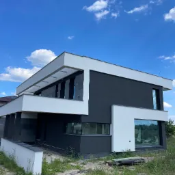 Nowoczesny dom z płaskim dachem, wykończony w kolorach bieli i grafitu, z dużymi oknami, na tle błękitnego nieba z chmurami. Teren wokół domu częściowo zagospodarowany, widoczny piasek i rosnąca...