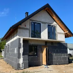 Dom w trakcie ocieplania: dolna część ścian pokryta szarymi płytami izolacyjnymi, górna część z białych pustaków, czarne okna, drewniane elementy dachu widoczne, błękitne niebo w tle.