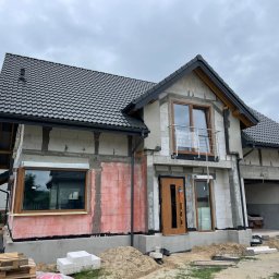 BANDINVESTMENT - Dom jednorodzinny w trakcie ocieplania elewacji, widoczne bloczki z betonu komórkowego, częściowo otynkowane, z zamontowanymi oknami i drzwiami, przygotowany pod ocieplenie styropianem, palety...