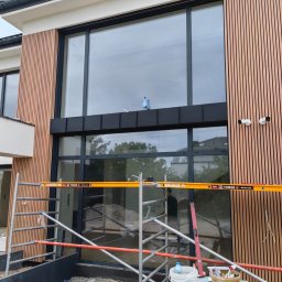 BAND INVESTMENT SPÓŁKA Z OGRANICZONĄ ODPOWIEDZIALNOŚCIĄ - Nowoczesna elewacja domu z drewnianymi panelami pionowymi i dużymi oknami, widoczne rusztowanie i akcesoria malarskie, sugerujące prace wykończeniowe.