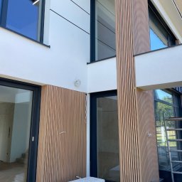 BAND INVESTMENT SPÓŁKA Z OGRANICZONĄ ODPOWIEDZIALNOŚCIĄ - Nowoczesna elewacja domu z drewnianymi panelami pionowymi i białym tynkiem. Widoczne okna z czarnymi ramami i fragment rusztowania. Częściowo ukończony montaż.