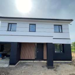 BAND INVESTMENT SPÓŁKA Z OGRANICZONĄ ODPOWIEDZIALNOŚCIĄ - Dwukondygnacyjny dom z nowoczesną elewacją: białe ściany z poziomymi pasami, czarne ceglane filary i drewniane panele przy wejściu. Widoczne okna i fragment dachu.