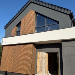 BANDINVESTMENT - Nowoczesny dom z drewnianą elewacją w pionowe panele i grafitowym tynkiem. Widoczne trójkątne okno i otwarte drewniane drzwi wejściowe. Osoba w oknie.