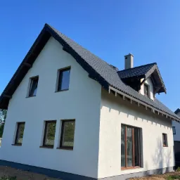 Biały dom jednorodzinny z szarą dachówką i kominem, widok z zewnątrz, elewacja po tynkowaniu, brązowe ramy okienne, błękitne niebo w tle.