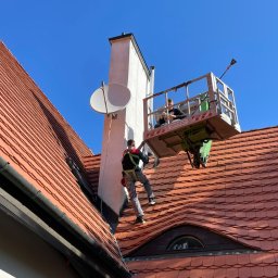 BANDINVESTMENT - Dwóch pracowników na podnośniku koszowym, jeden na dachu, zabezpieczeni uprzężami, pracują przy kominie na tle czerwonej dachówki i błękitnego nieba.