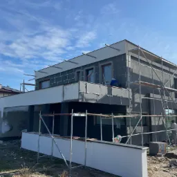 Nowoczesny dom w trakcie ocieplania, widoczne rusztowania, styropianowe płyty elewacyjne i pracownicy na tle błękitnego nieba.