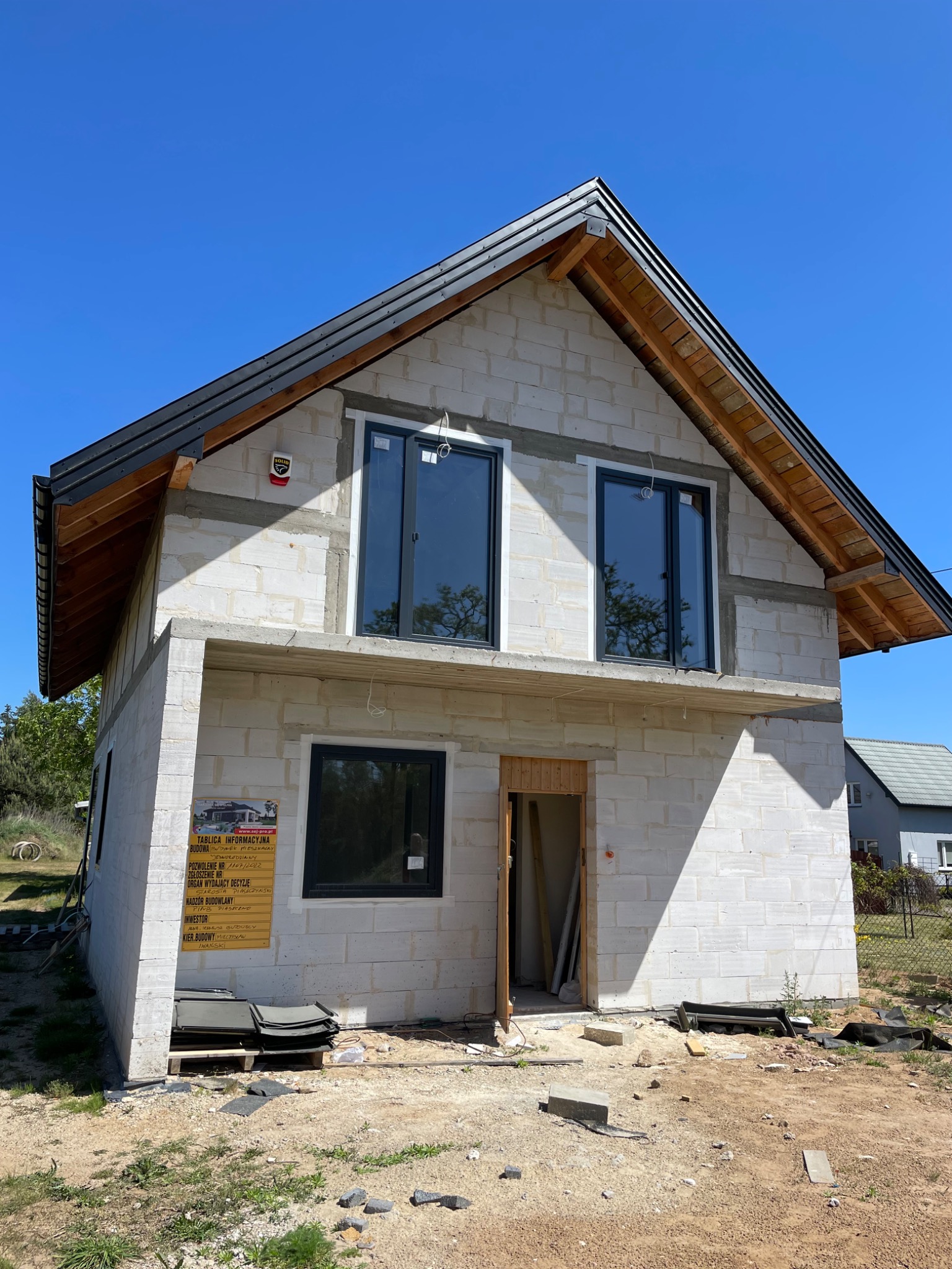 Surowa elewacja dwukondygnacyjnego domu z pustaków, widoczne czarne ramy okienne i częściowo zamontowane drewniane elementy dachu, tablica informacyjna budowy z boku budynku.