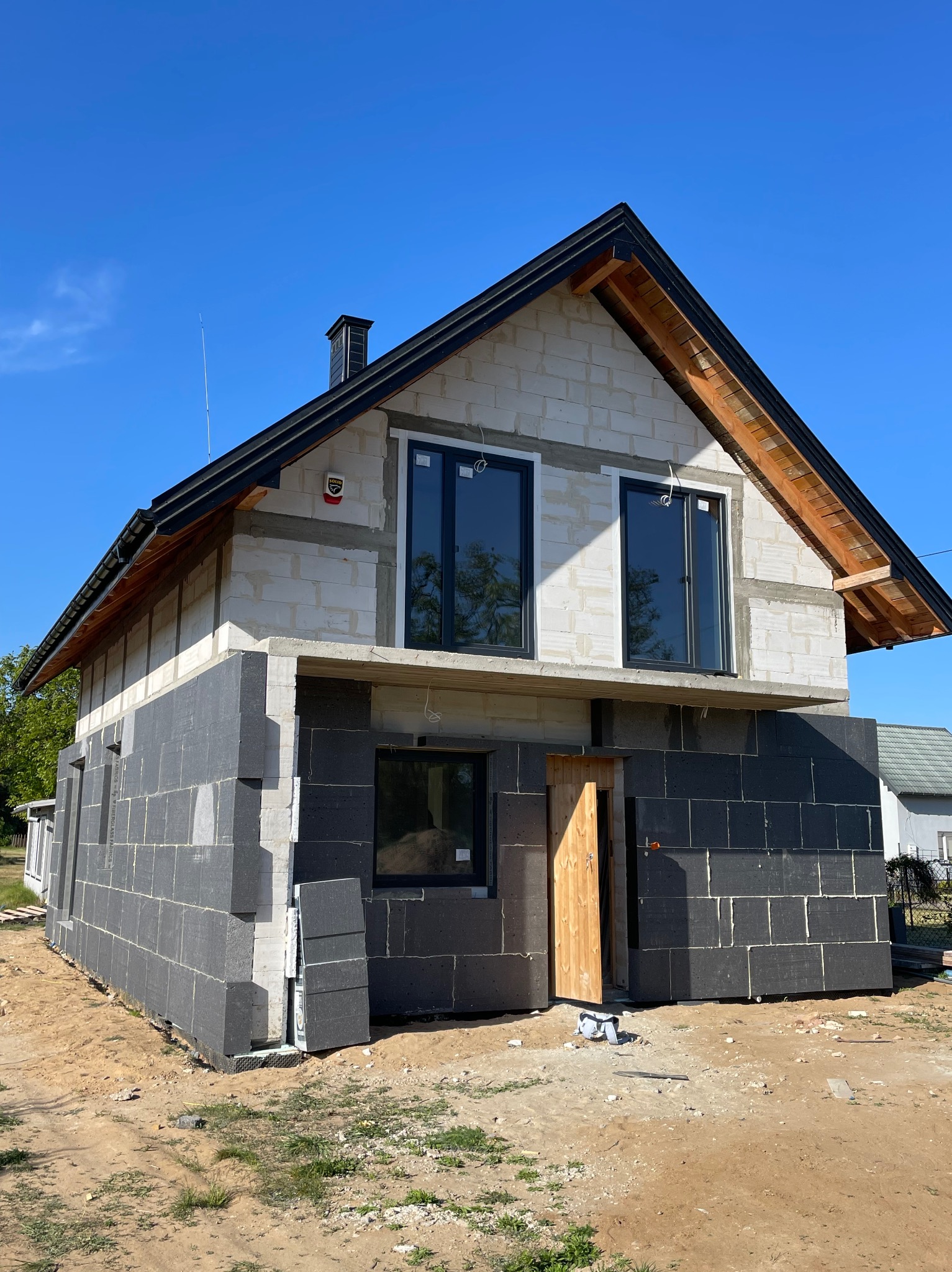 Dom w trakcie ocieplania: dolna część ścian pokryta szarymi płytami izolacyjnymi, górna część z białych pustaków, czarne okna, drewniane elementy dachu widoczne, błękitne niebo w tle.