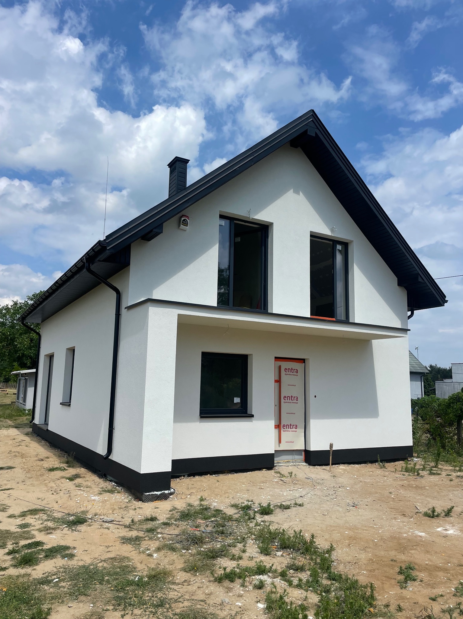 Biały dom jednorodzinny z czarnym dachem i orynnowaniem, świeżo otynkowany, z widocznymi zabezpieczeniami drzwi wejściowych, na działce z piaskiem i skąpą roślinnością.