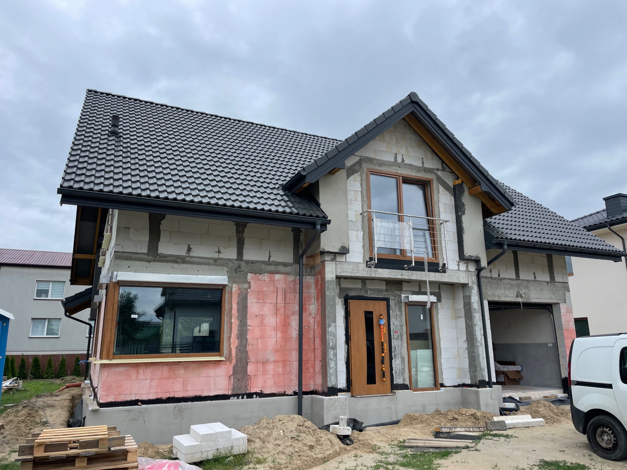 Dom jednorodzinny w trakcie ocieplania elewacji, widoczne bloczki z betonu komórkowego, częściowo otynkowane, z zamontowanymi oknami i drzwiami, przygotowany pod ocieplenie styropianem, palety...