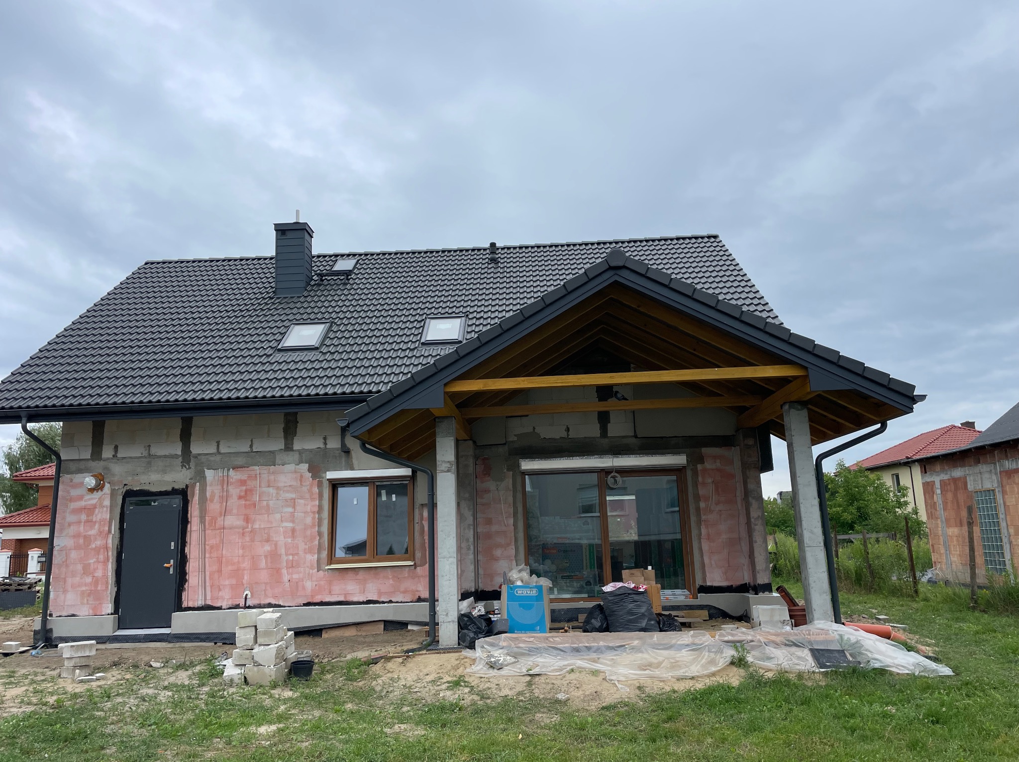 Niedokończony dom jednorodzinny z widocznymi pustakami, częściowo otynkowaną elewacją i zamontowaną stolarką okienną i drzwiową; dach pokryty szarą dachówką ceramiczną, widoczne elementy...