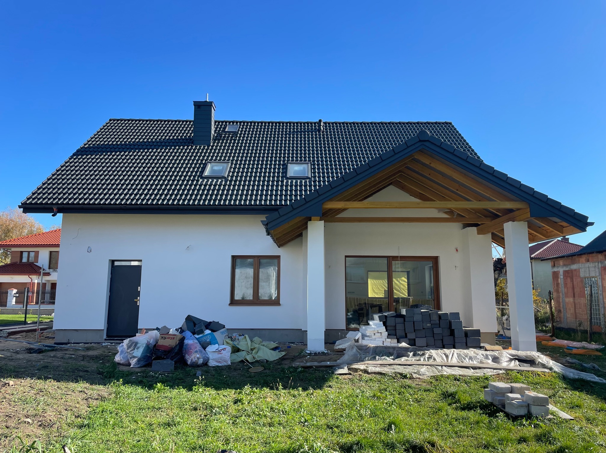 Nowoczesny dom jednorodzinny z białymi ścianami i ciemną dachówką, widoczne stosy kostki brukowej i worki z materiałami budowlanymi przed budynkiem, sugerujące prace wykończeniowe.