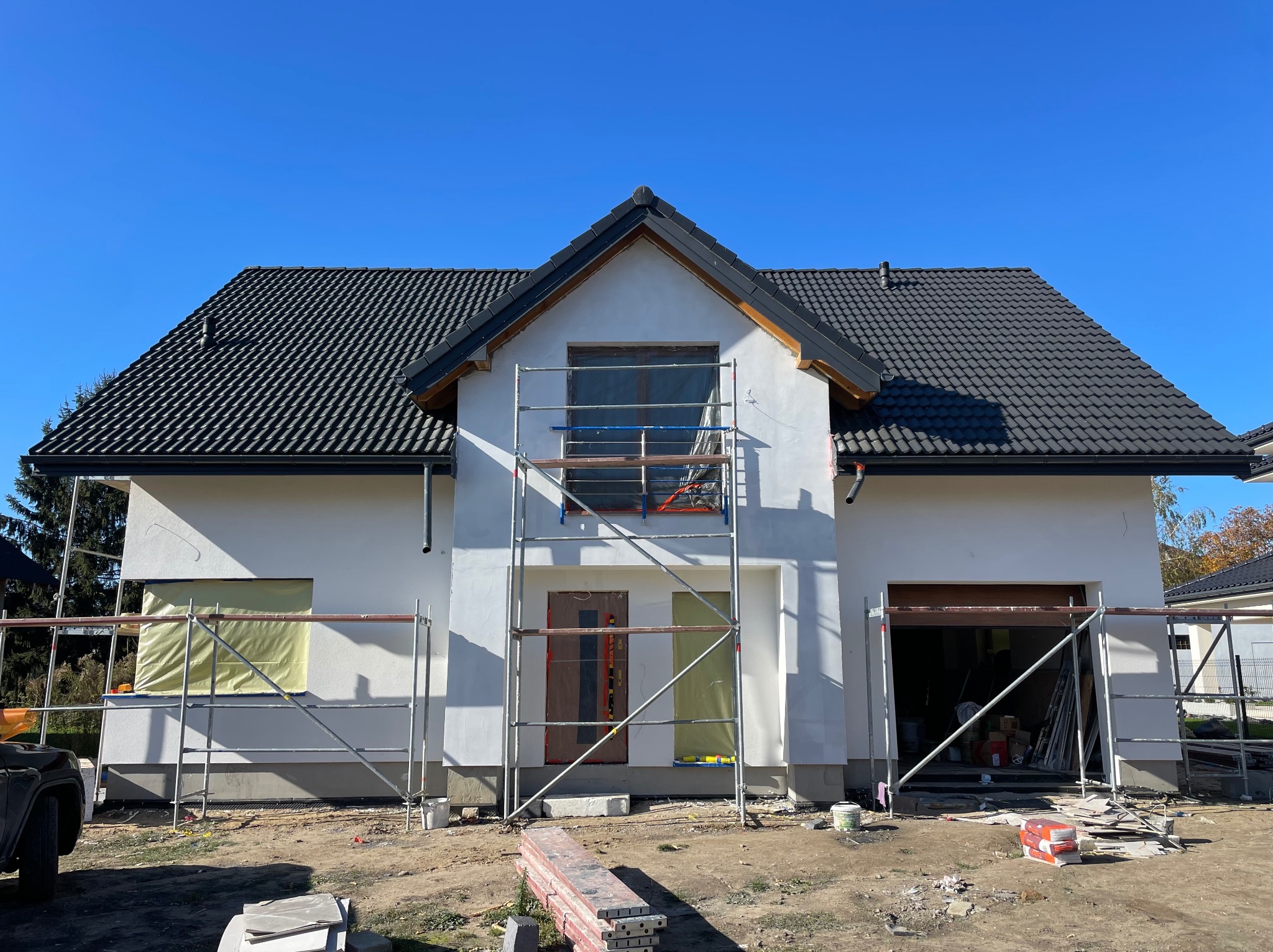 Dwukondygnacyjny dom z ciemną dachówką, w trakcie prac elewacyjnych, z widocznym rusztowaniem, okna i drzwi zabezpieczone folią, teren wokół domu częściowo uporządkowany, błękitne niebo.