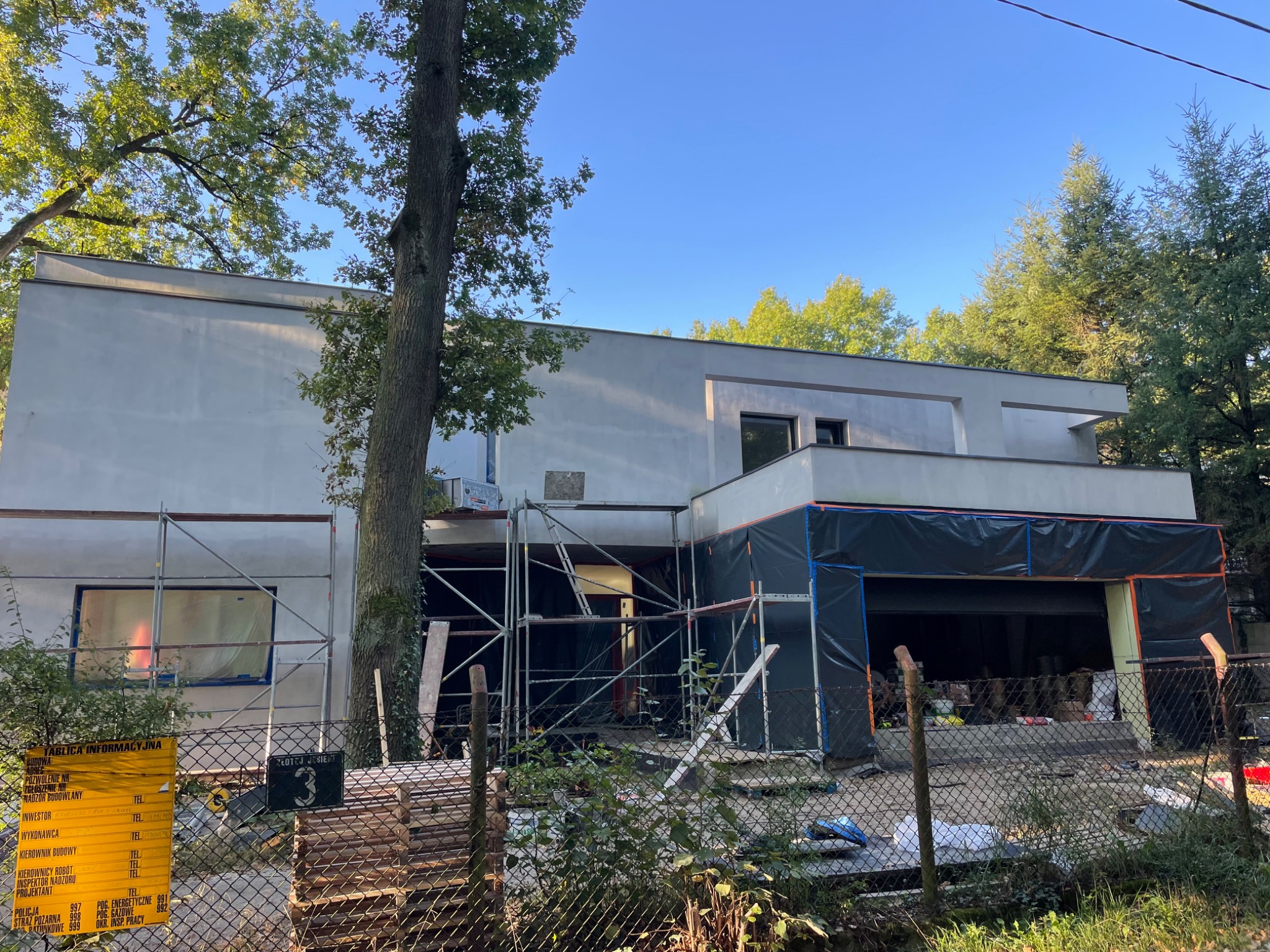 Nowoczesny dom w trakcie wykończenia elewacji, widoczne rusztowania i zabezpieczenia folią, tablica informacyjna budowy przy ogrodzeniu.