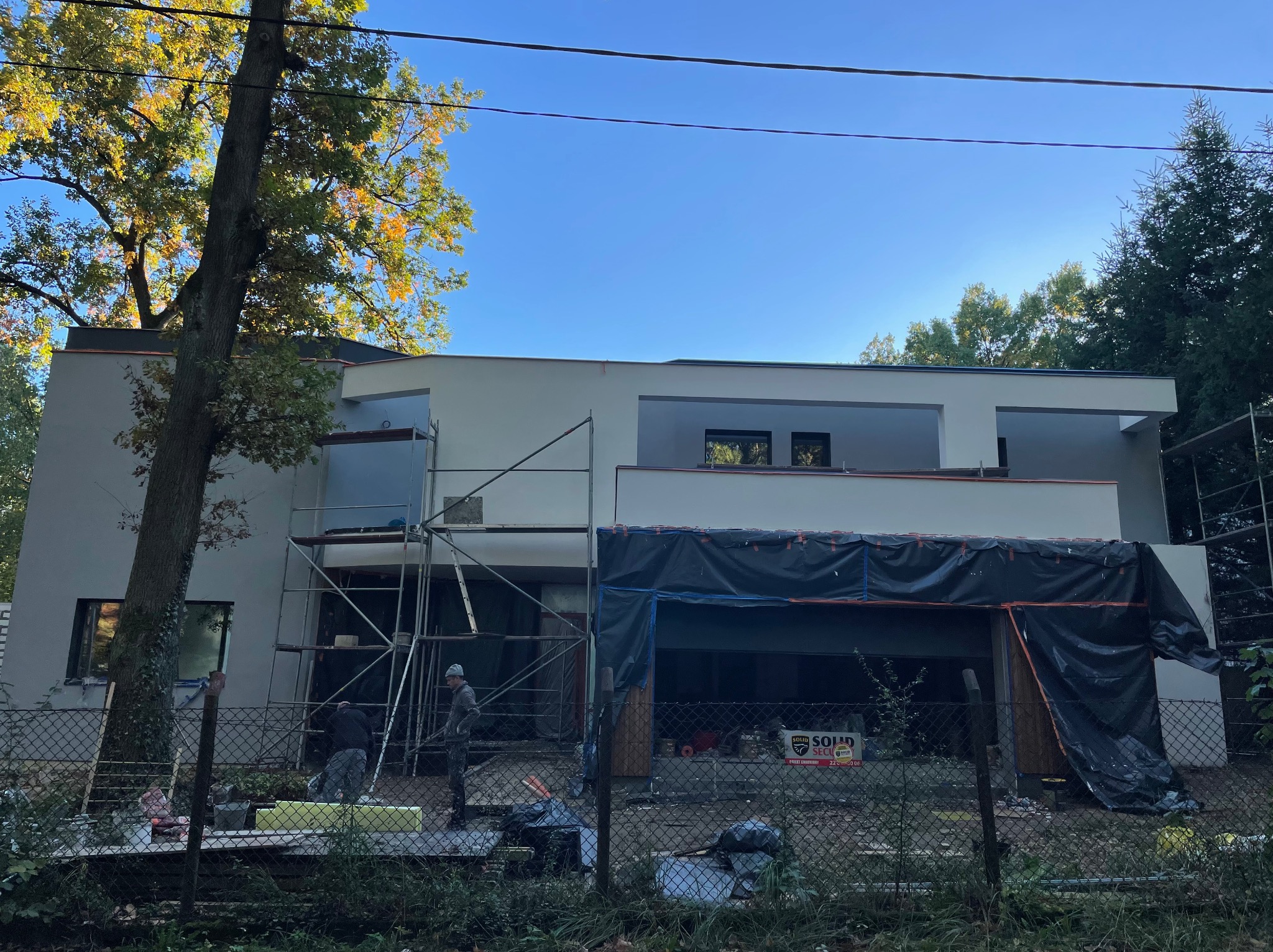 Nowoczesny dom w trakcie prac elewacyjnych: szare tynki, rusztowania, robotnicy przy pracy, widoczne materiały izolacyjne i zabezpieczenia folią, drzewo z jesiennymi liśćmi w tle.