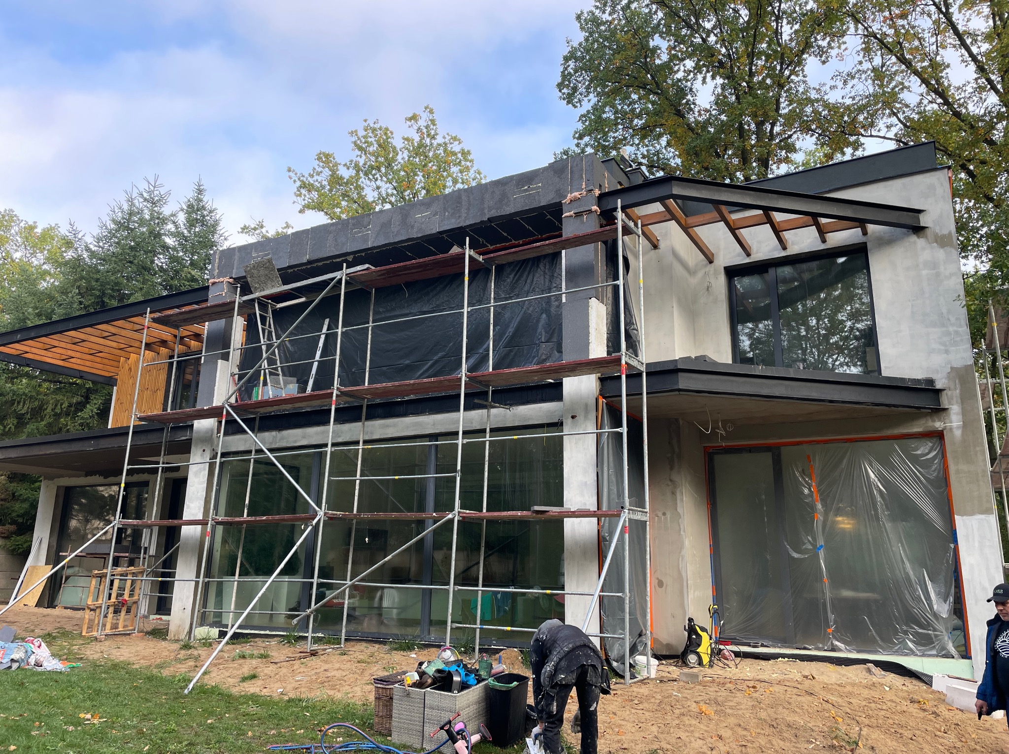 Dwukondygnacyjny dom w trakcie tynkowania elewacji, widoczne rusztowania, zabezpieczone okna folią, pracownik przygotowujący zaprawę, częściowo ocieplona ściana szarą wełną mineralną.