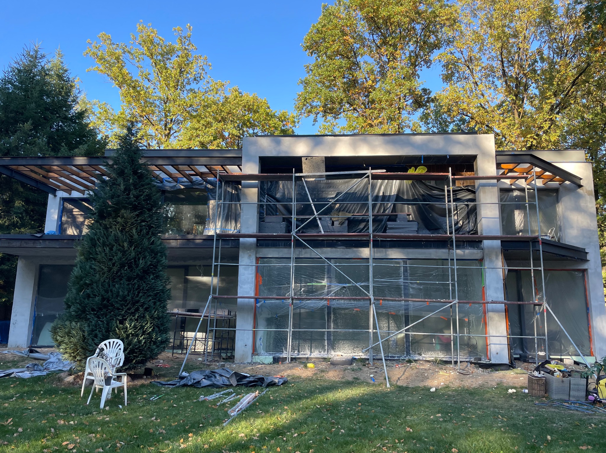 Nowoczesny dom w trakcie tynkowania elewacji, zabezpieczony folią i rusztowaniem, z widocznymi elementami konstrukcji dachu i otaczającą zielenią.