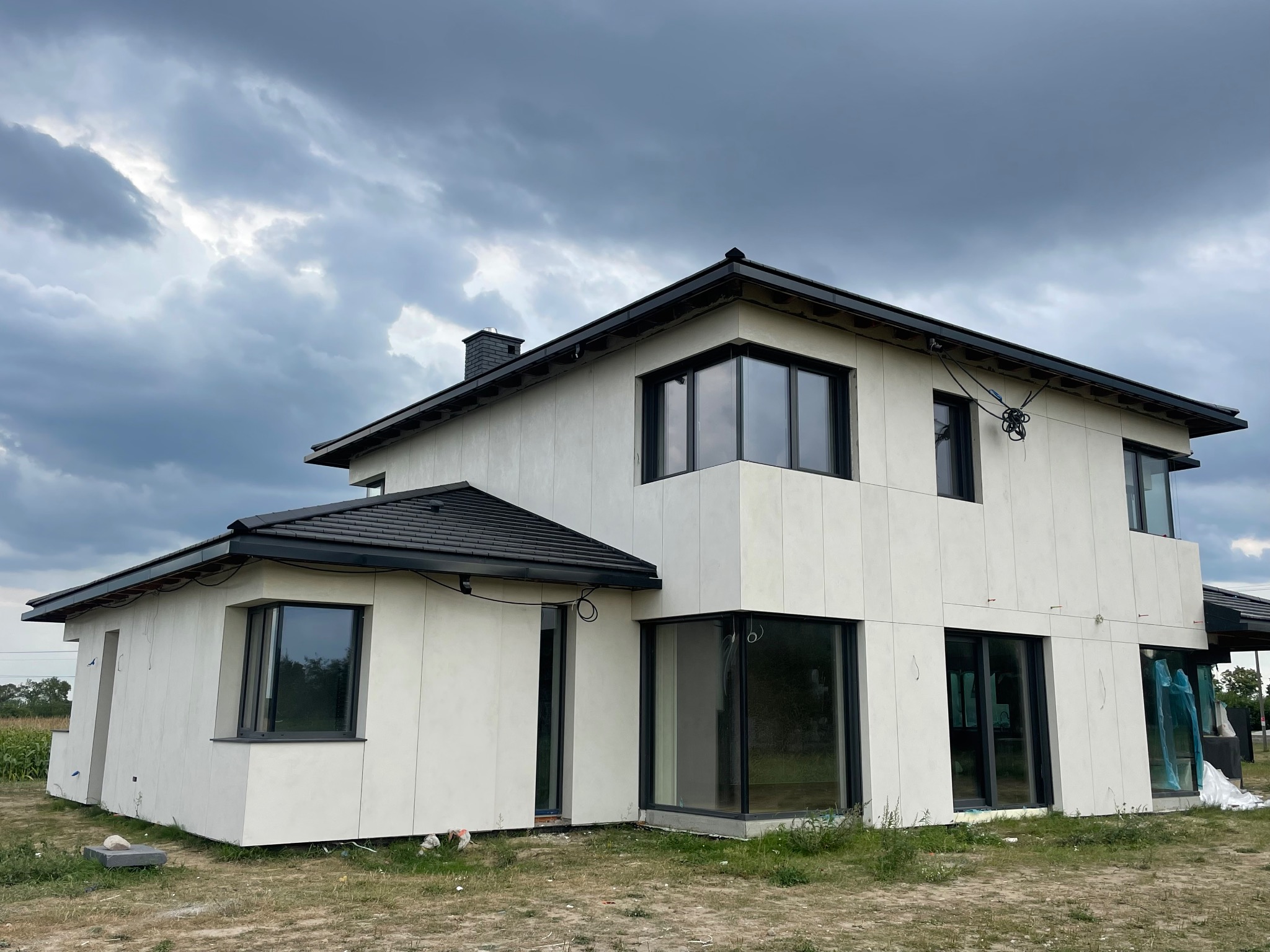 Nowoczesny dom dwukondygnacyjny z elewacją w jasnych panelach, czarną stolarką okienną i dachem. Widoczne elementy instalacji na zewnątrz. Pochmurne niebo w tle.