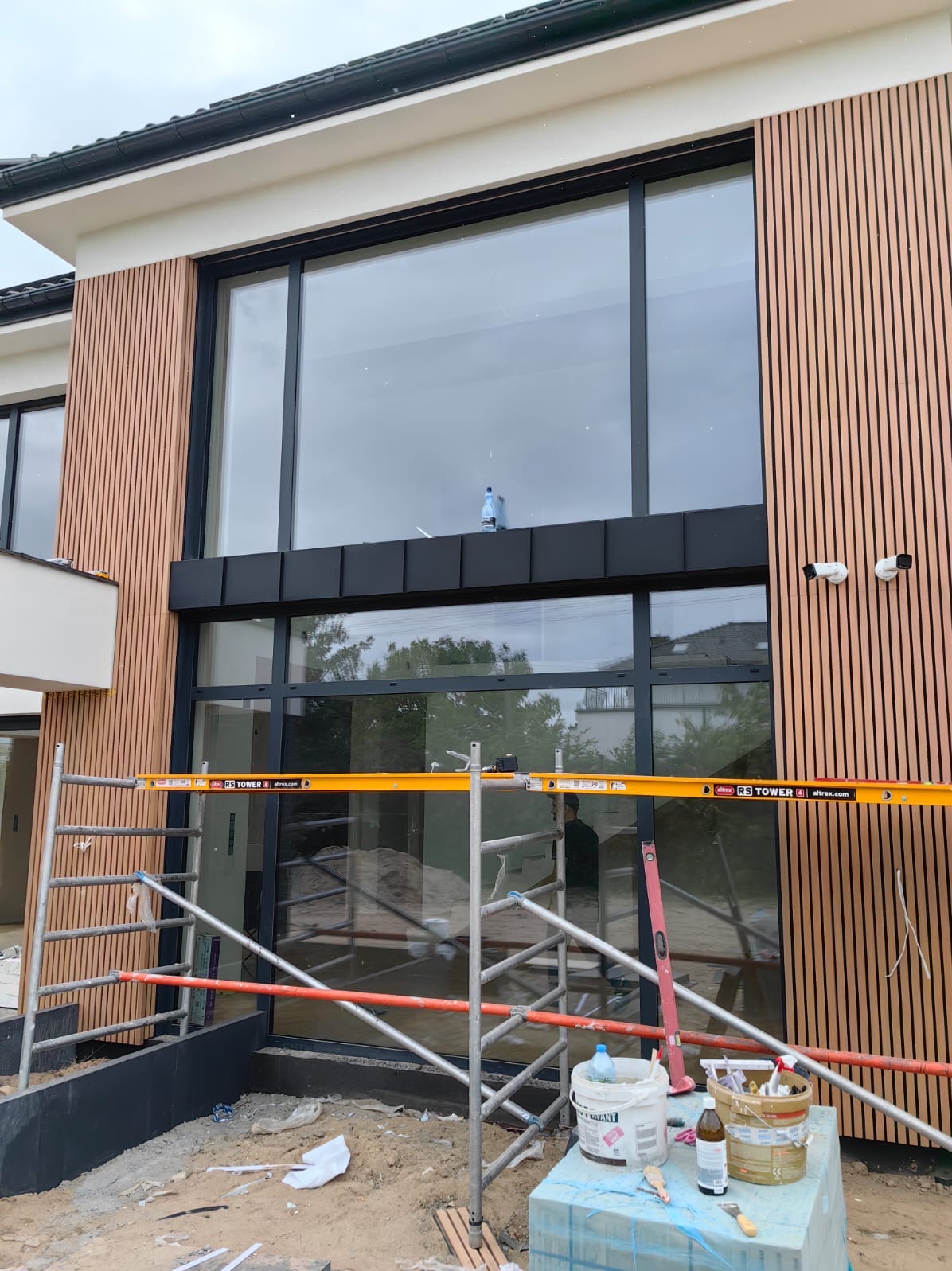 Nowoczesna elewacja domu z drewnianymi panelami pionowymi i dużymi oknami, widoczne rusztowanie i akcesoria malarskie, sugerujące prace wykończeniowe.