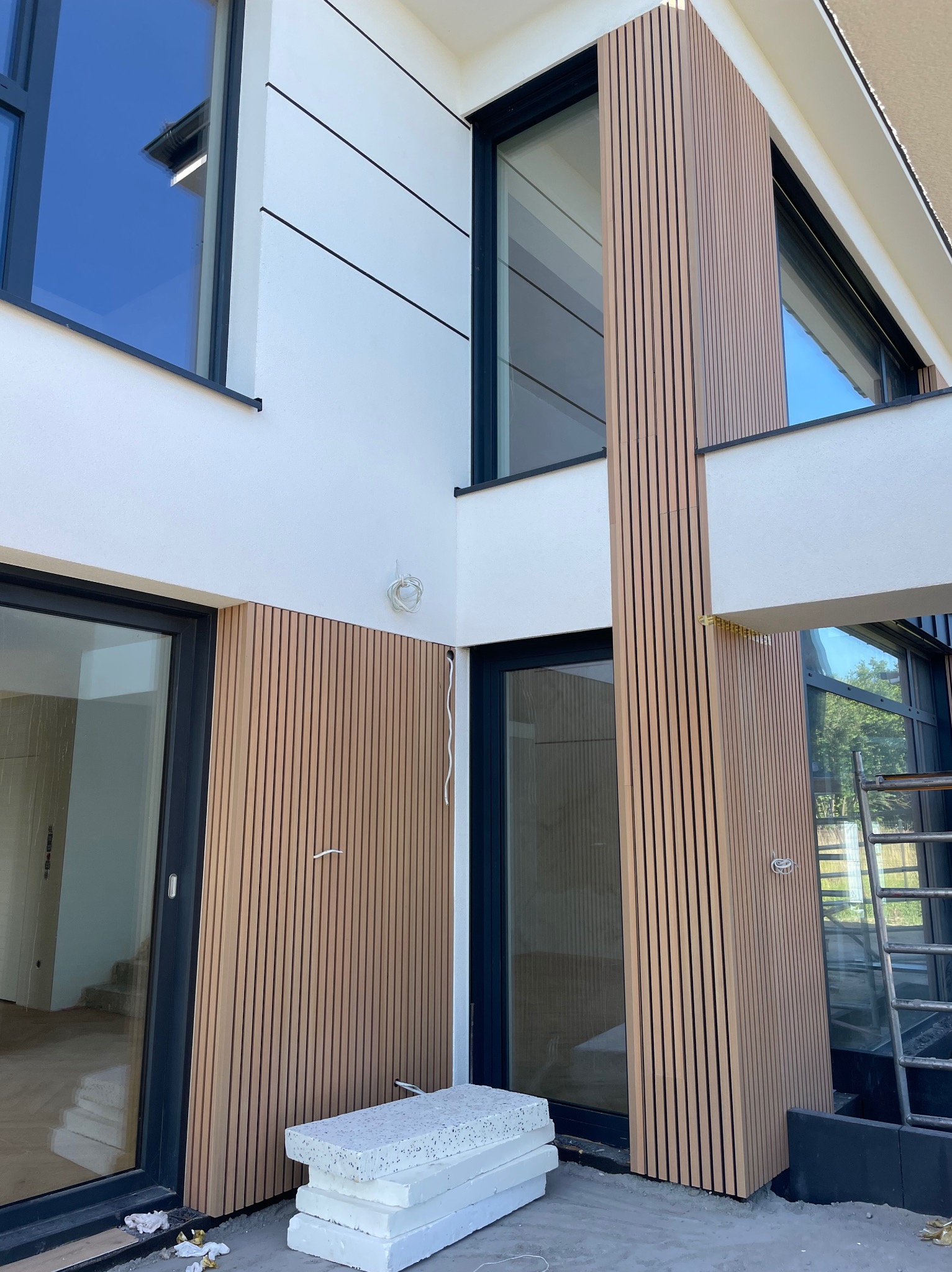 Nowoczesna elewacja domu z drewnianymi panelami pionowymi i białym tynkiem. Widoczne okna z czarnymi ramami i fragment rusztowania. Częściowo ukończony montaż.