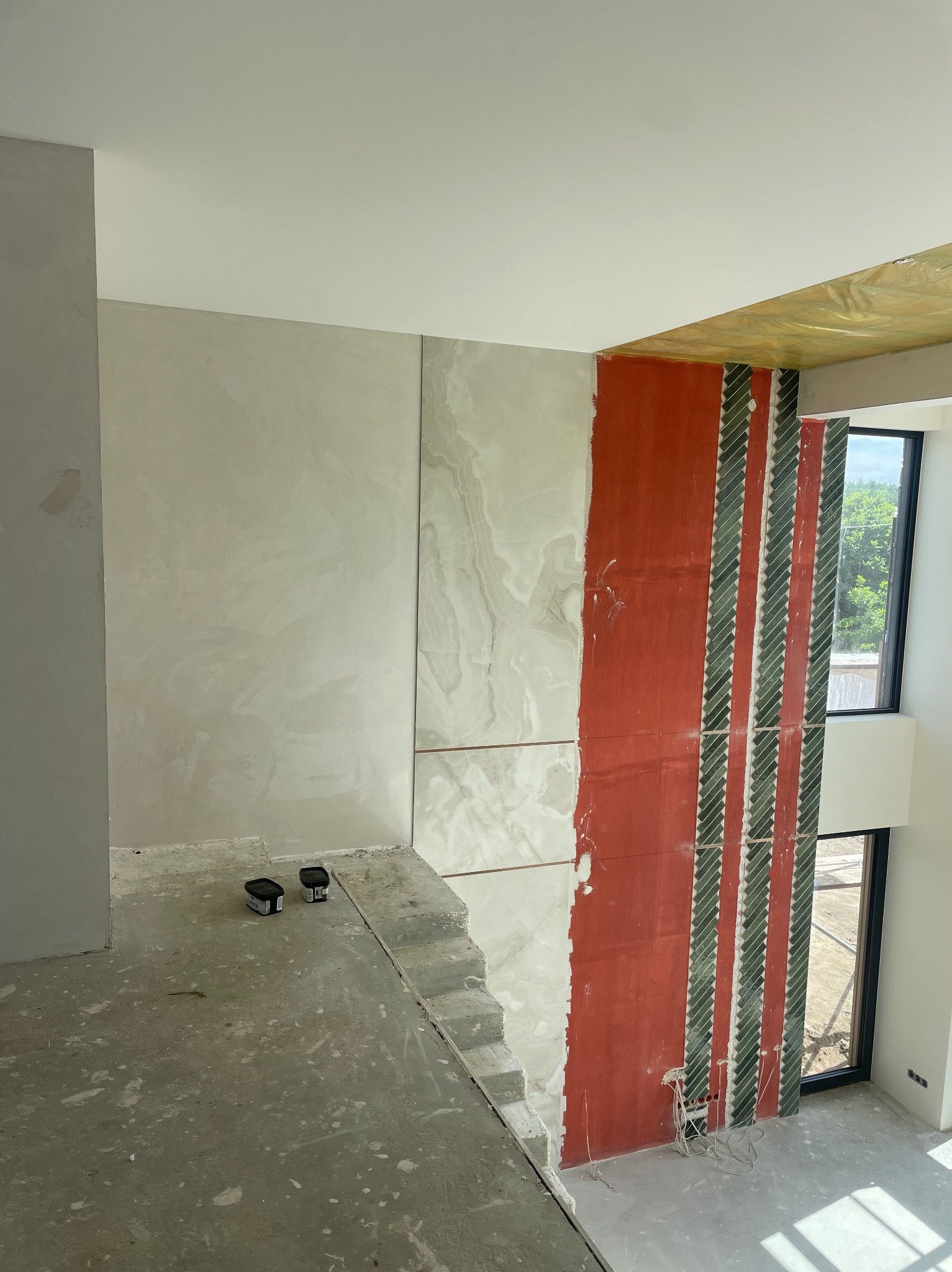 Wnętrze w trakcie wykańczania: fragment ściany z dekoracyjnym tynkiem, płytkami imitującymi marmur i pionowymi pasami z zielonej mozaiki. Widoczne puszki z farbą.