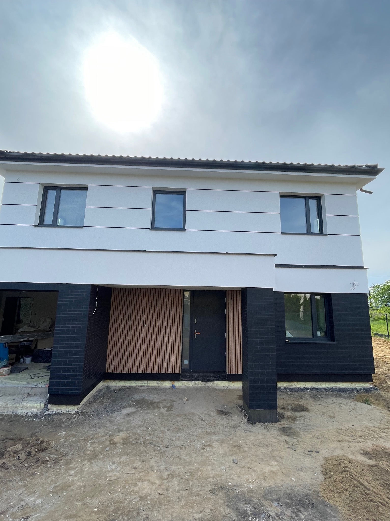 Dwukondygnacyjny dom z nowoczesną elewacją: białe ściany z poziomymi pasami, czarne ceglane filary i drewniane panele przy wejściu. Widoczne okna i fragment dachu.