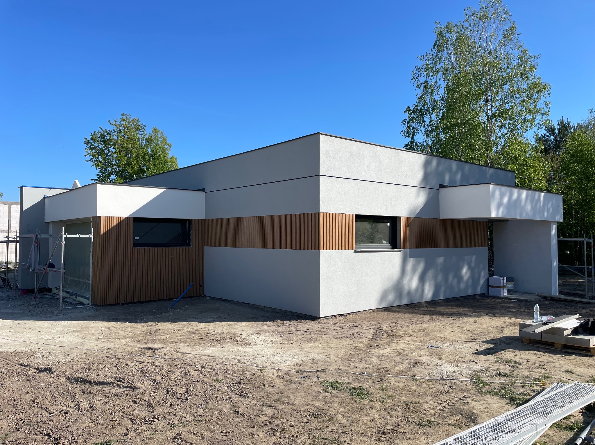 Nowoczesny dom z szarą elewacją i drewnianymi panelami, okna z czarnymi ramami, błękitne niebo w tle. Częściowo widoczne rusztowanie.