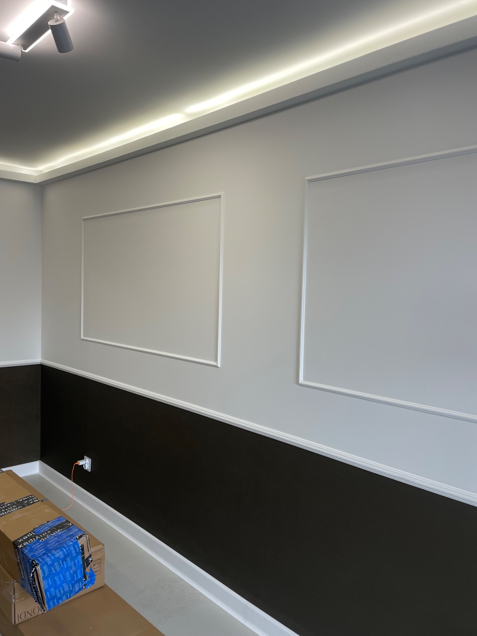 Dwukolorowa ściana z białymi listwami dekoracyjnymi i oświetleniem LED podsufitowym. Dolna część ściany ciemnobrązowa, górna jasnoszara. W rogu kartony.