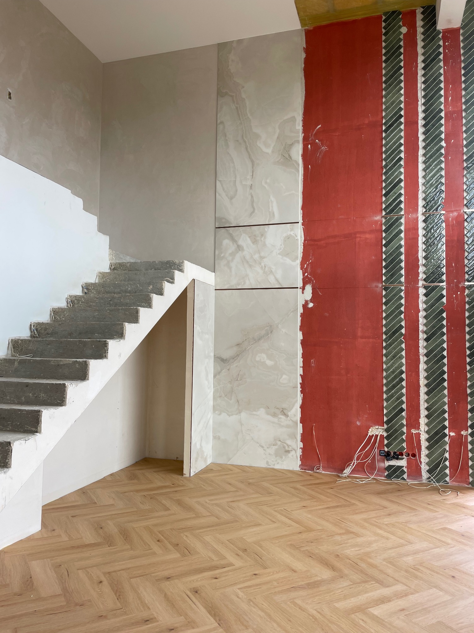 Wnętrze w trakcie wykończenia: surowe schody, fragment ściany z płytami imitującymi marmur, druga ściana z częściowo ułożoną mozaiką i widoczną instalacją elektryczną.