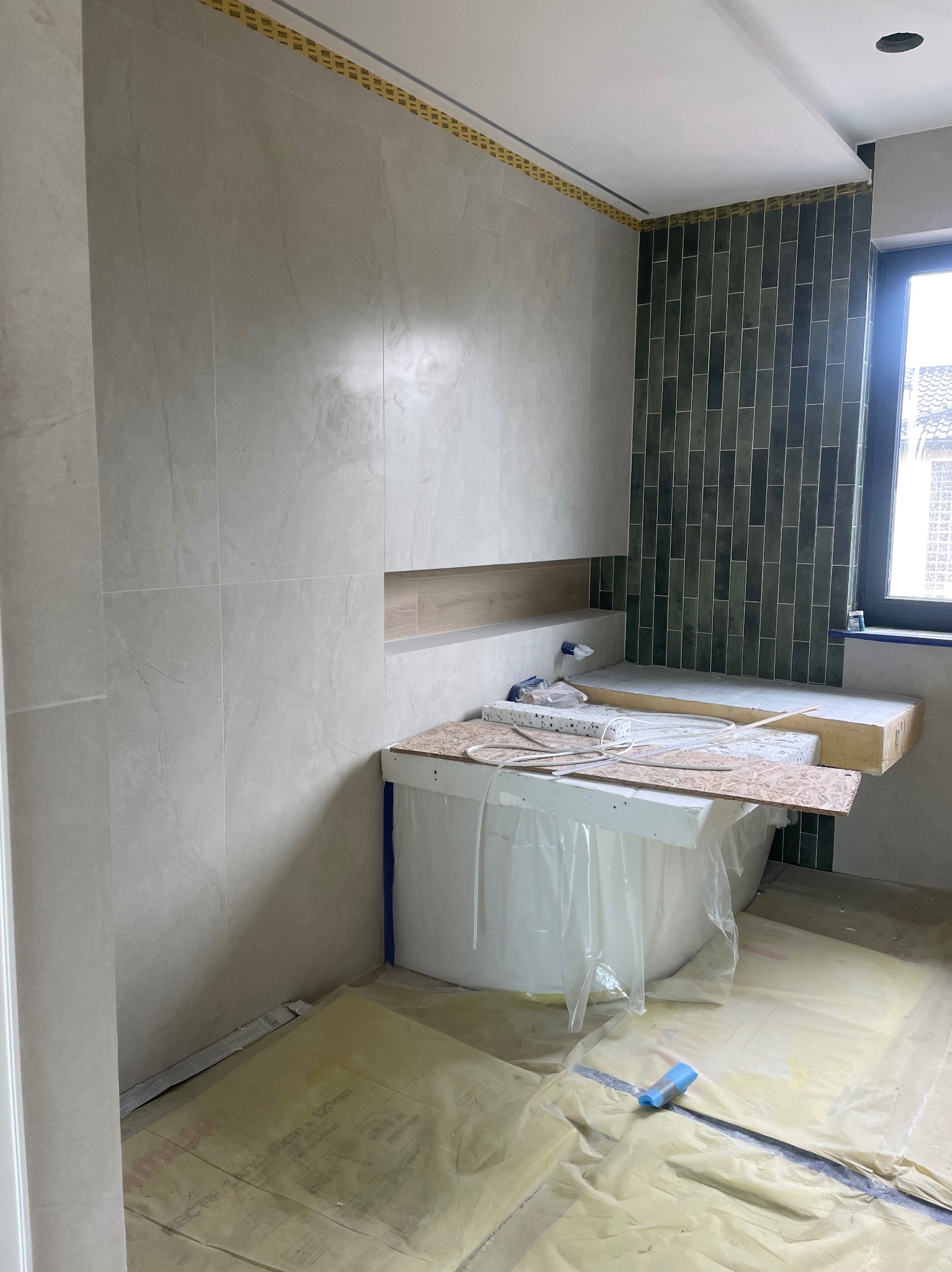 Wnętrze łazienki w trakcie wykańczania: jasne płytki na ścianach, ciemnozielona mozaika, zabezpieczona folią wanna i podłoga. Widoczne materiały budowlane.