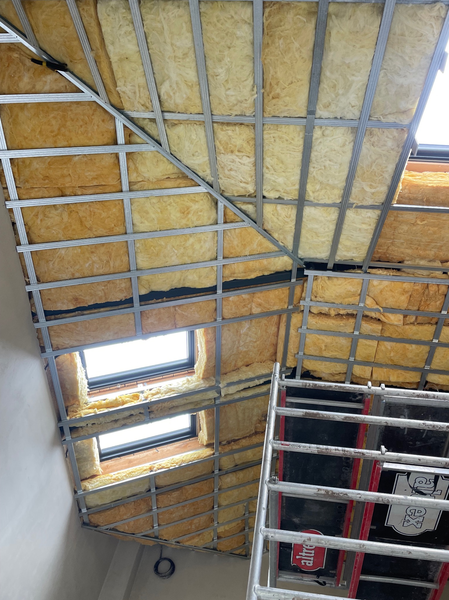 Skośny sufit poddasza z oknami połaciowymi w trakcie ocieplania wełną mineralną, widoczna metalowa konstrukcja nośna i fragment rusztowania.