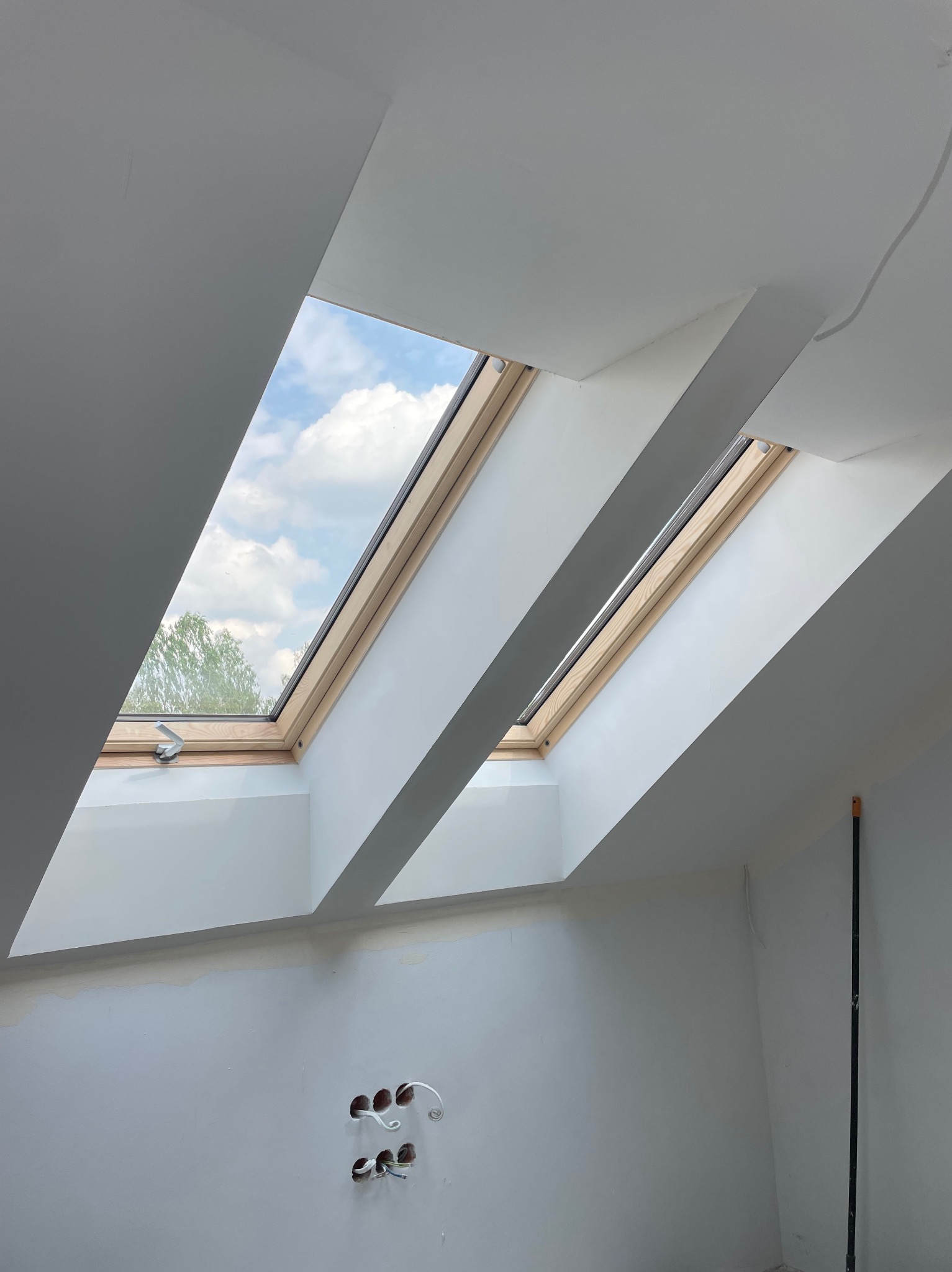 Białe wnętrze poddasza z dwoma oknami dachowymi, widoczne ślady po gładzi na ścianach i wystające kable elektryczne. W tle błękitne niebo.