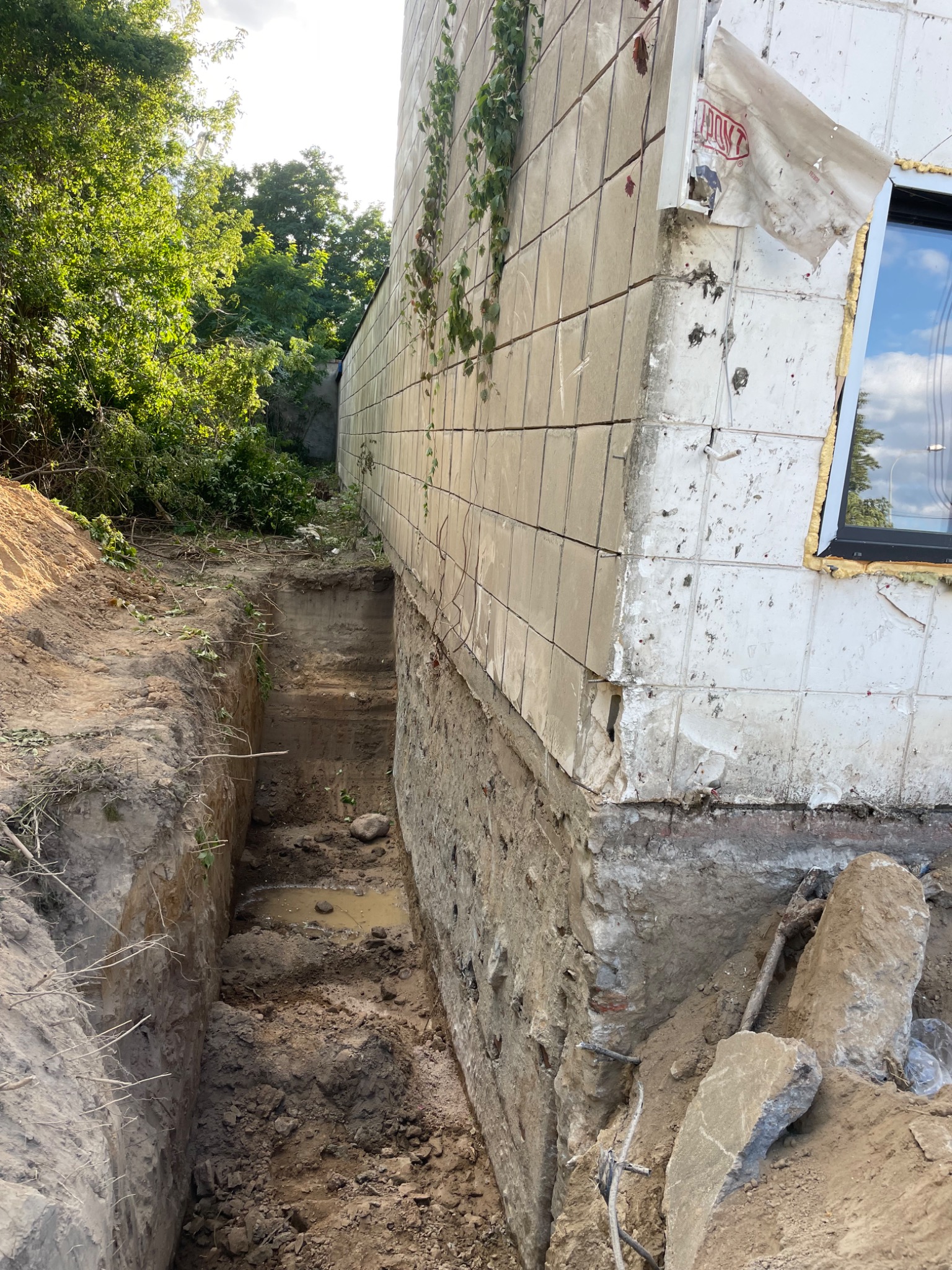 Odkopany fundament budynku z uszkodzoną elewacją z płytek, widoczne ślady demontażu. W tle zieleń drzew. Prace przygotowawcze do renowacji.
