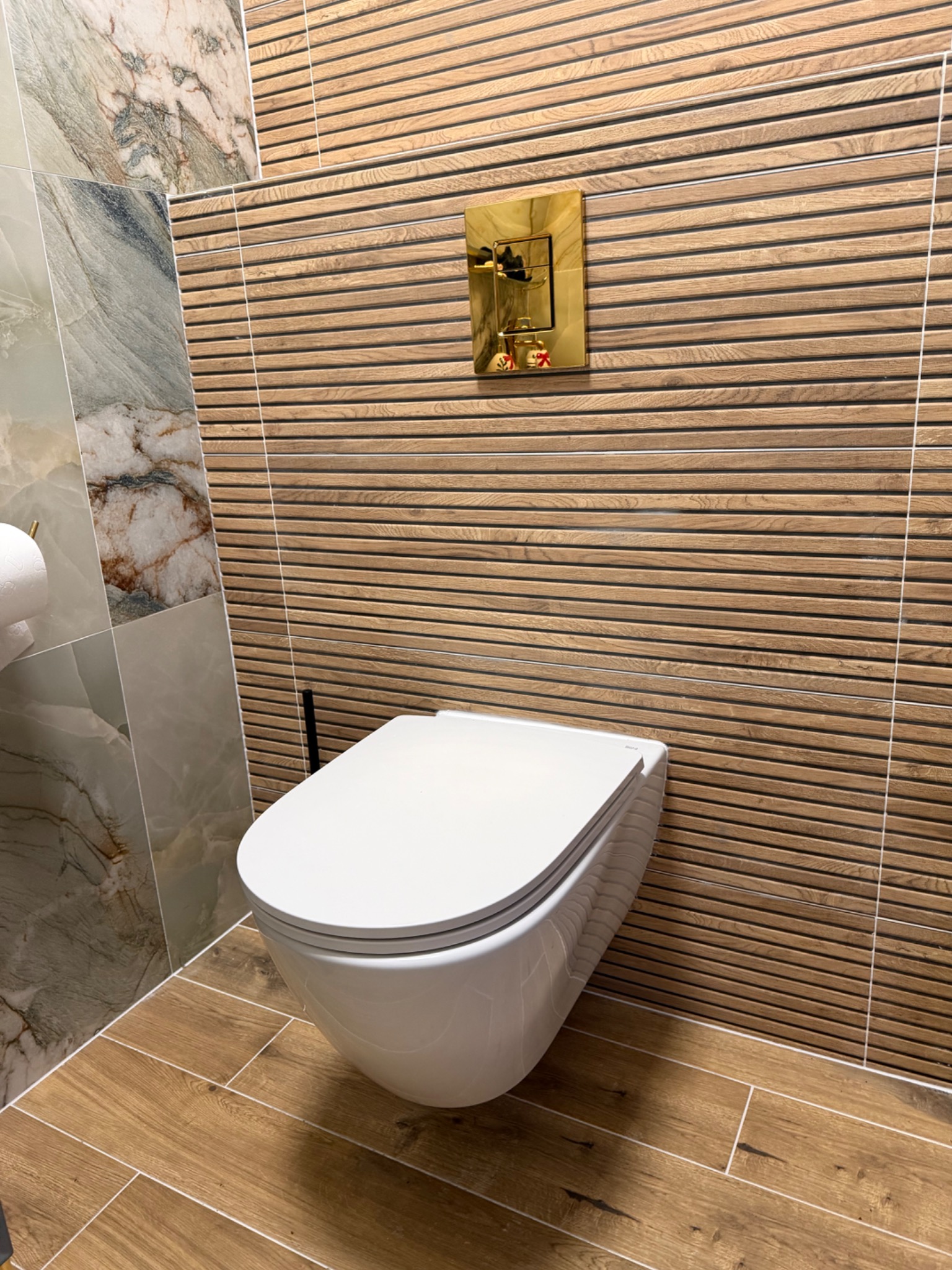 Nowoczesna toaleta podwieszana w Mrokowie. Drewnopodobne płytki na ścianie i podłodze, marmurowe akcenty. Złota armatura dodaje elegancji.
