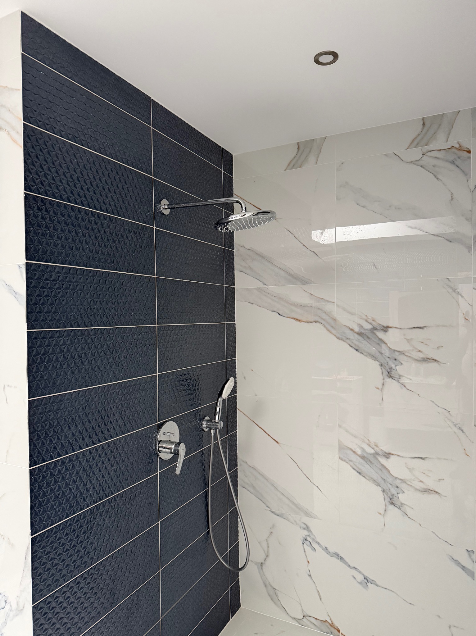 Wykończona kabina prysznicowa z czarnymi, trójwymiarowymi płytkami i białymi płytkami imitującymi marmur. Widoczna bateria i deszczownica.