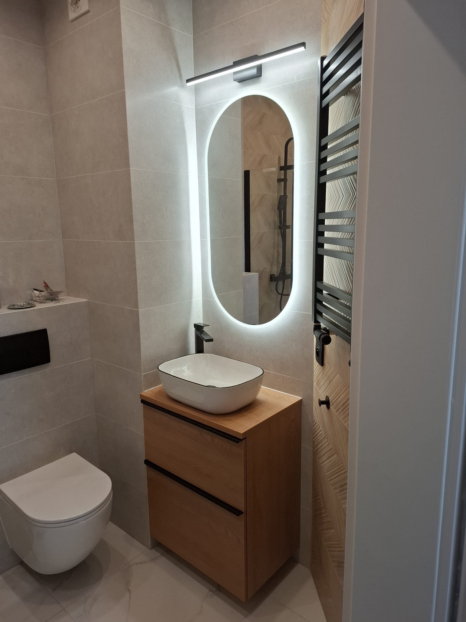 Nowoczesna łazienka z owalnym lustrem LED, drewnianą szafką pod umywalką i grzejnikiem drabinkowym na tle płytek w odcieniach szarości i beżu.