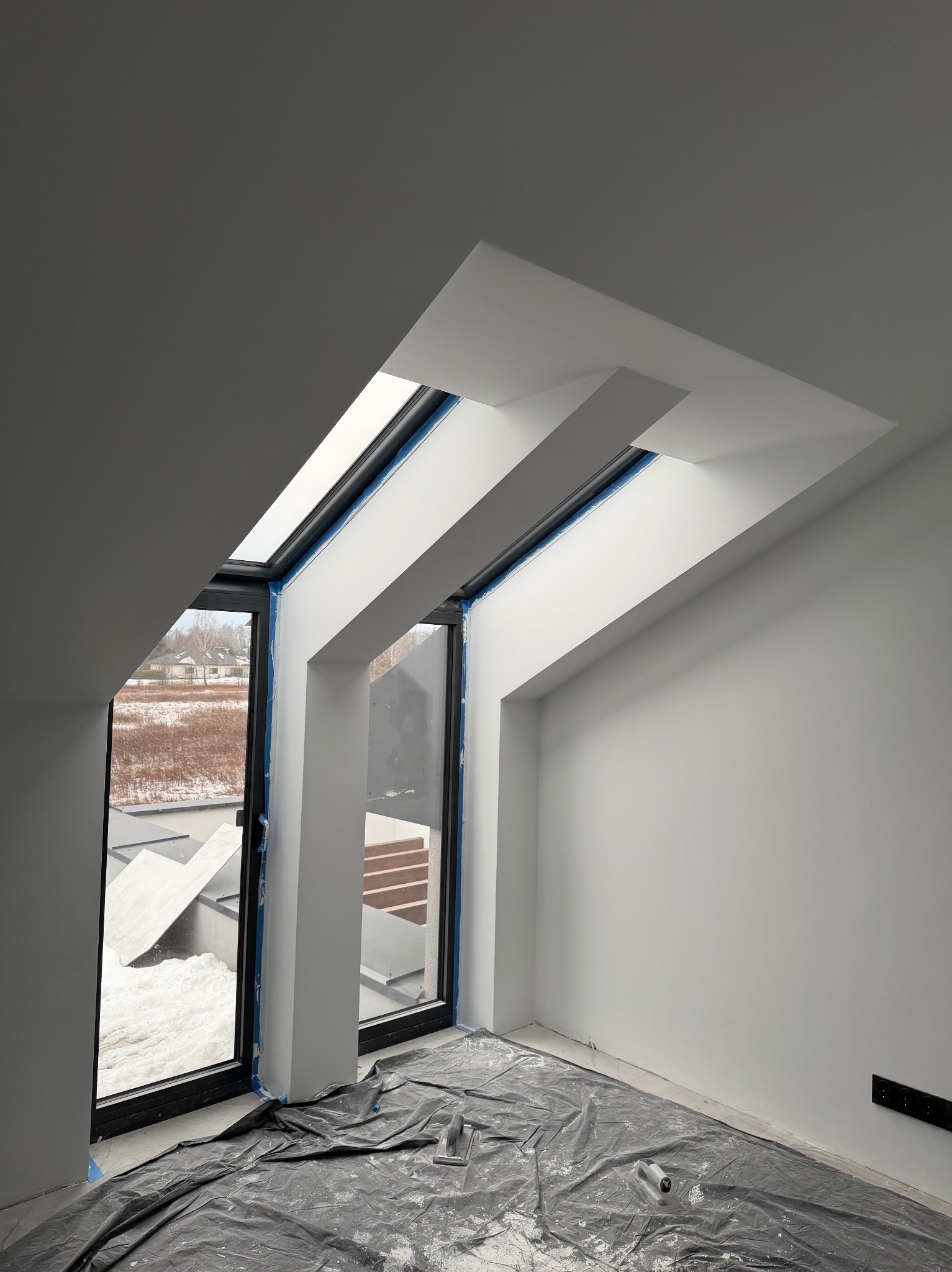 Poddasze ze skośnym oknem dachowym po tynkowaniu, zabezpieczone folią malarską. Widoczne narzędzia i świeżo otynkowane ściany w odcieniach bieli i szarości.