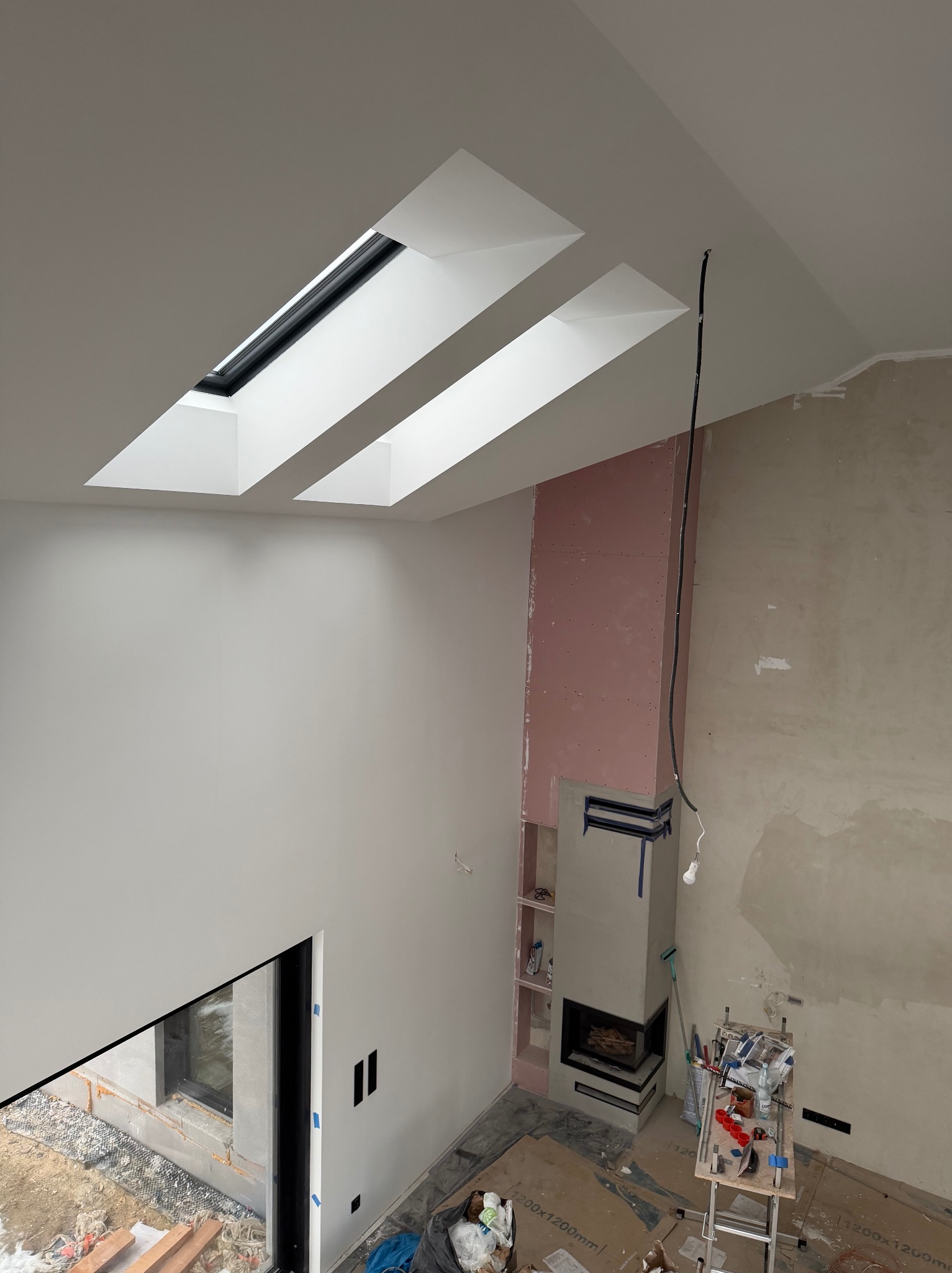 Wnętrze w trakcie wykończenia: ściany przygotowane do malowania, kominek obłożony płytami gipsowymi, widoczne okna dachowe i elementy instalacji elektrycznej.