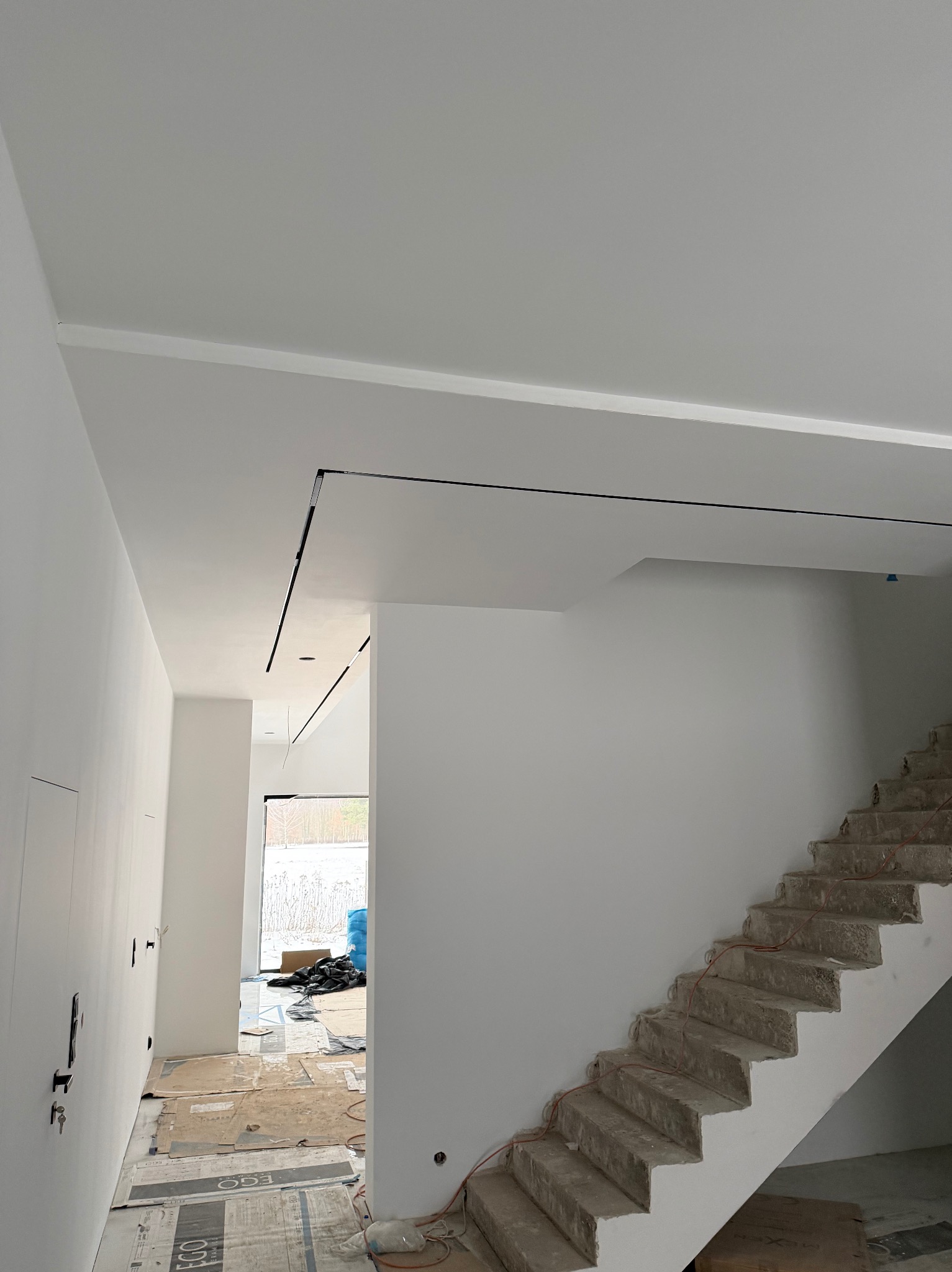 Wnętrze w trakcie wykańczania: białe ściany, sufit z czarnymi listwami, surowe schody betonowe, zabezpieczona podłoga. Widok na okno i materiały budowlane.