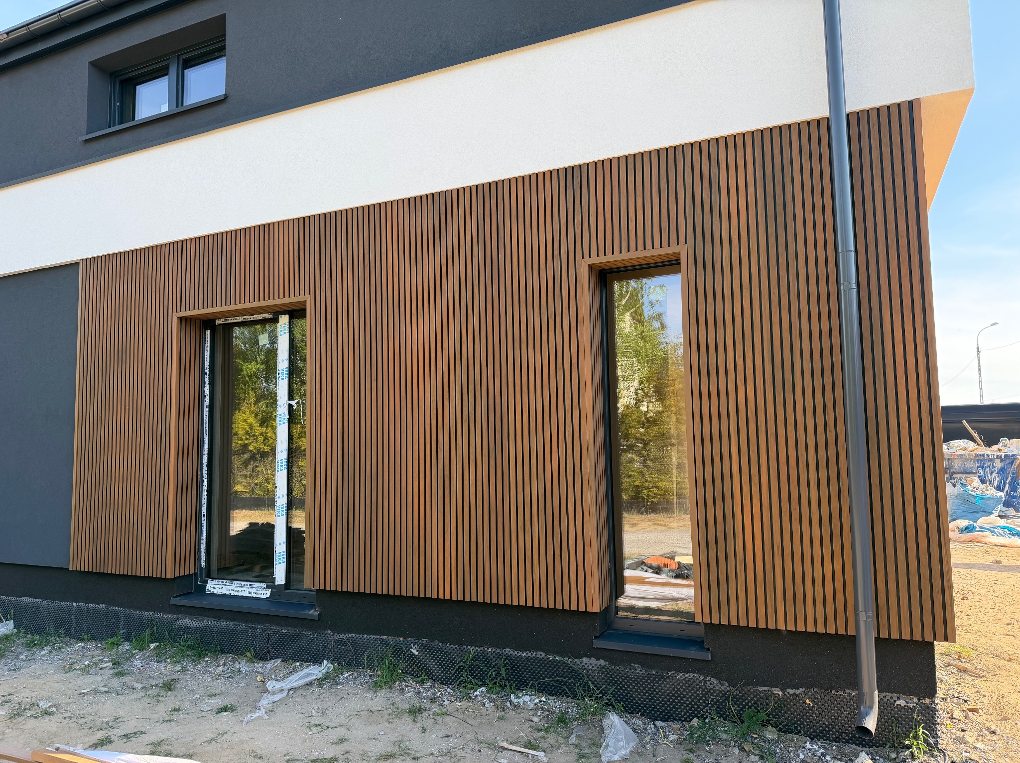 Nowoczesna elewacja domu z drewnianymi panelami pionowymi, szarą górną częścią i białym pasem. Widoczne okna z refleksami otoczenia i fragment rynny.