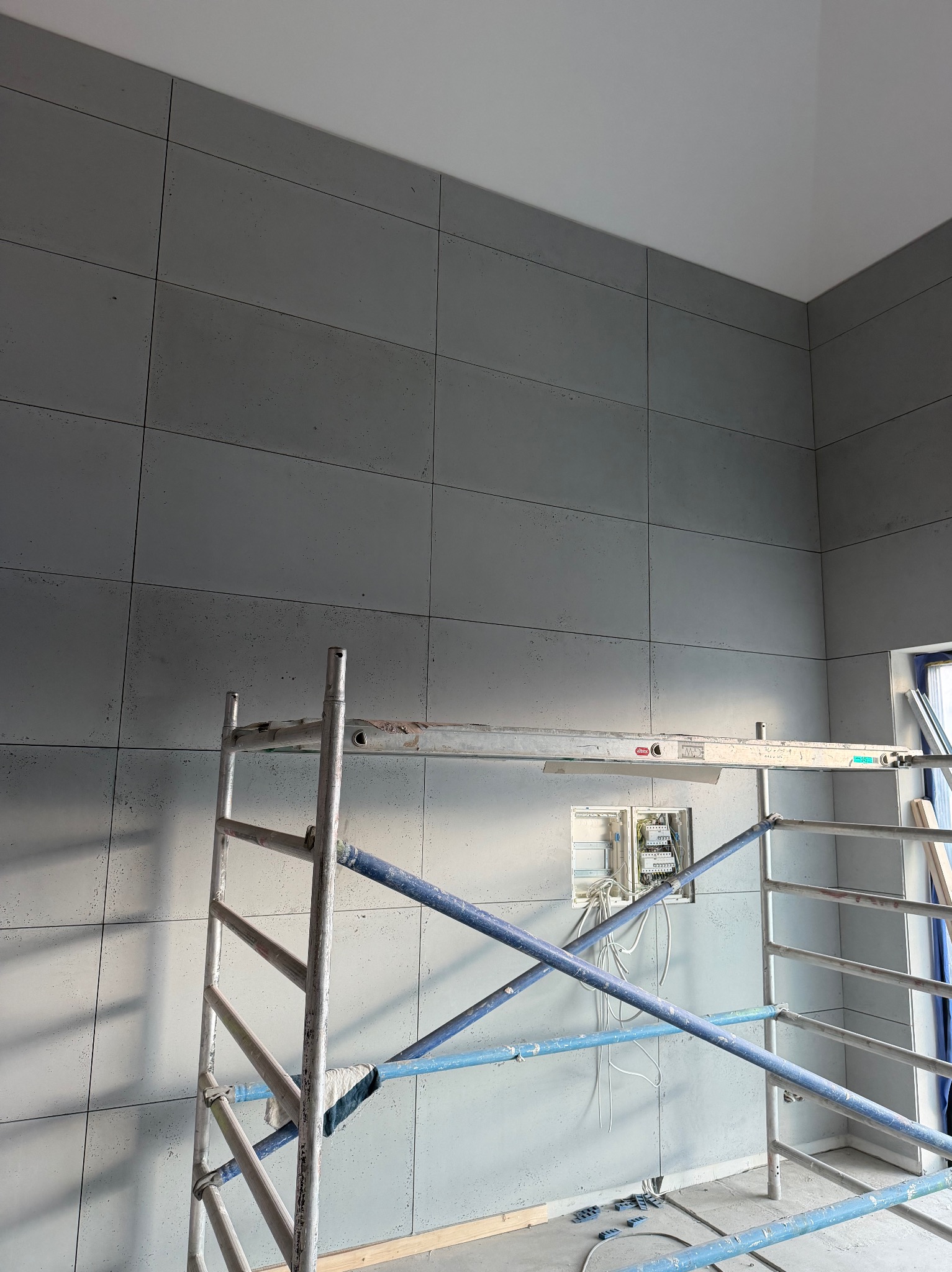 Ściana wykończona szarymi płytami imitującymi beton, widoczne rusztowanie i skrzynka elektryczna. Minimalistyczny design wnętrza w trakcie prac wykończeniowych.