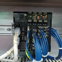 Panel sterowania automatyki przemysłowej Omron NX-ECC201 z podłączonymi niebieskimi kablami, widoczne diody LED sygnalizujące pracę modułów, białe kable Ethernet i oznaczenia na przewodach.